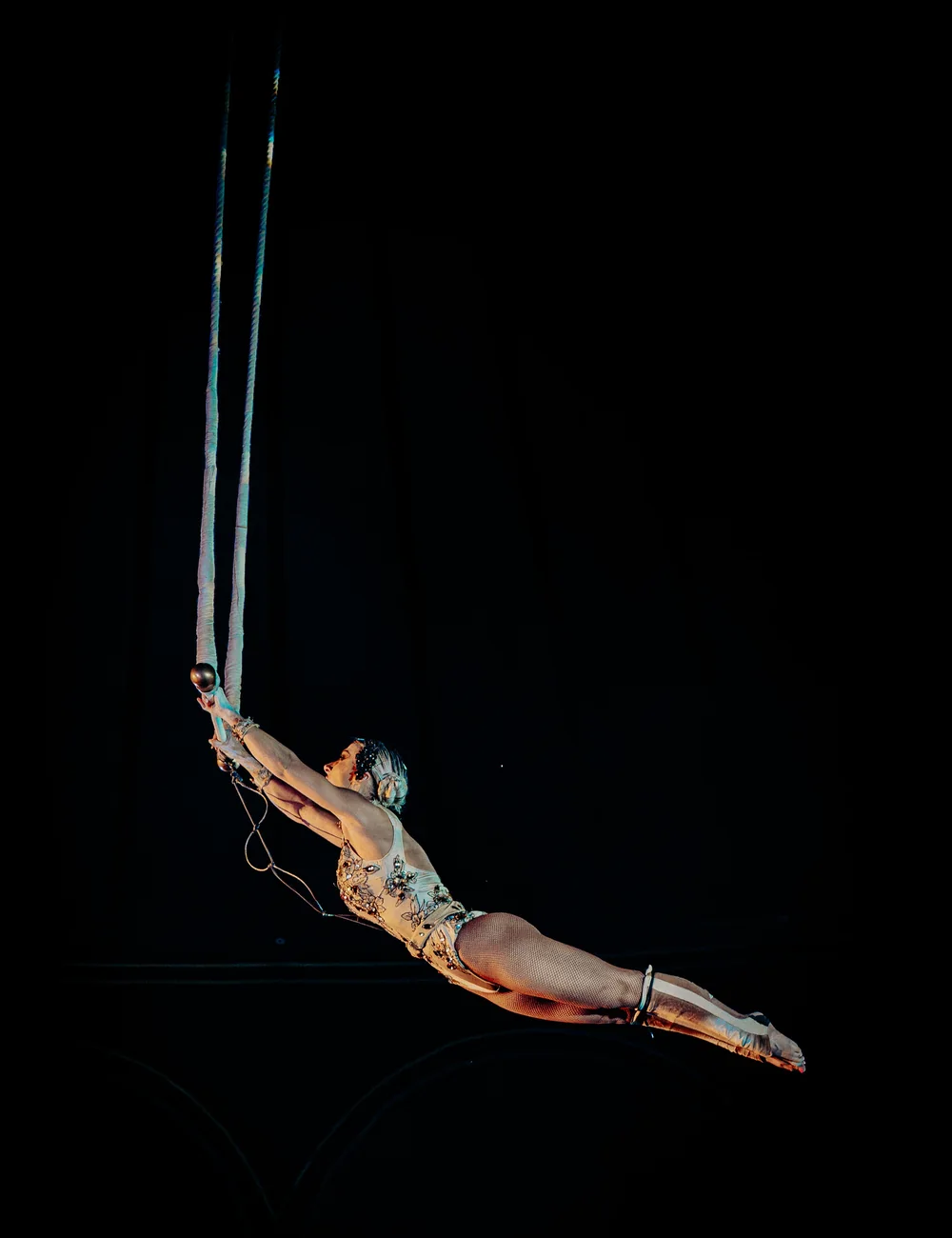 An aerial acrobat suspended on ropes, arched backward mid-performance against a dark background (representative photograph of an aerial act, not Maison Milano's own cast)